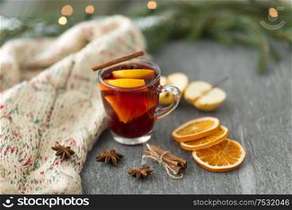 christmas and seasonal treats concept - glass of hot mulled wine with dry orange slicess and cinnamon, star anise, knitted scarf and apples on grey stone surface. glass of hot mulled wine with orange and cinnamon