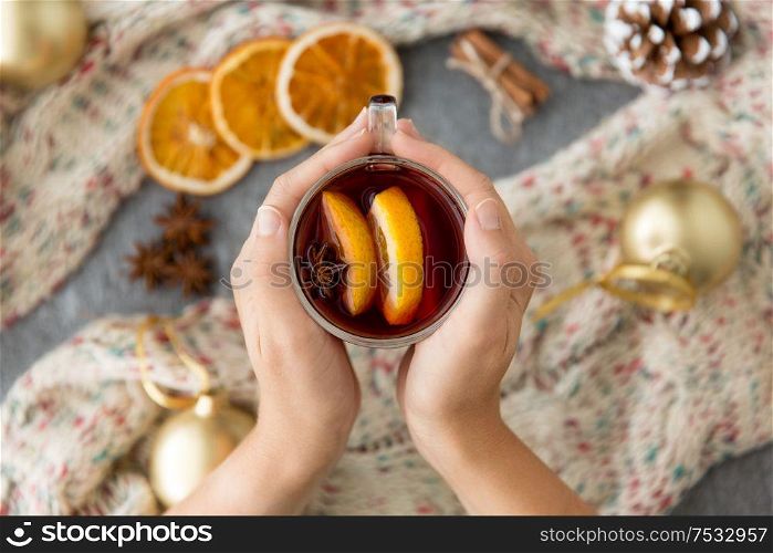 christmas and seasonal treats concept - woman&rsquo;s hands holding glass of hot mulled wine with orange slicess and star anise over knitted woolen scarf. hands with glass of hot mulled wine on christmas