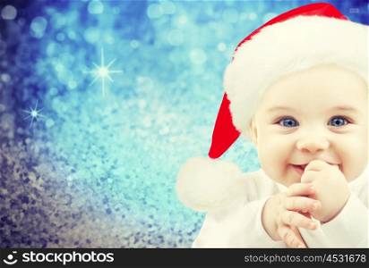 christmas, babyhood, childhood and people concept - happy baby in santa hat over blue glitter holidays lights background