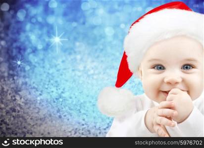 christmas, babyhood, childhood and people concept - happy baby in santa hat over blue glitter holidays lights background