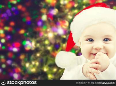 christmas, babyhood, childhood and people concept - happy baby in santa hat over holidays lights background