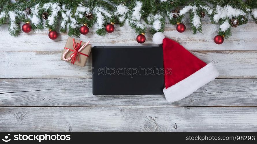 Christmas background with Santa Claus cap, electronic notebook and gift with traditional decorations on rustic white wood.