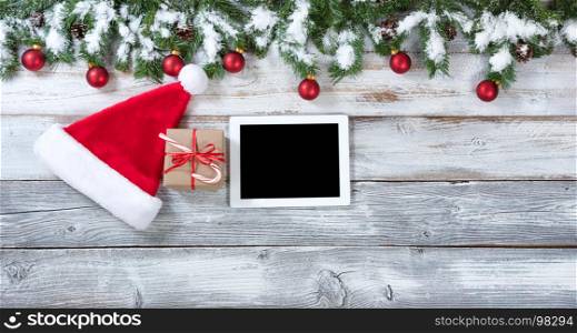 Christmas background with Santa Claus hat, electronic notebook and gift with traditional decorations on rustic white wood.