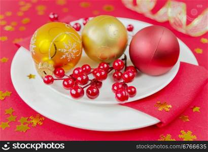 Christmas balls and holly berries are on the white porcelain plate, located on the red tablecloth strewn with golden confetti in the shape of snowflakes