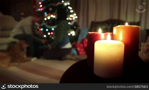 Christmas candles with a man, woman, and dog by the Christmas tree in the background. The woman unwraps a gift from the man, then hugs him.