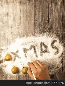 Christmas card with a female hand writing xmas, gold balls in the snow on a wooden background