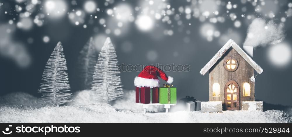Christmas card with cozy small decorative house , gifts and Santa Claus cap on sleigh on snow over fairy tale winter fir forest background and falling snow . New Year design postcard . Copy space for text. Christmas card with house in snow