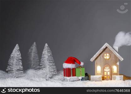 Christmas card with small glowing decorative house , gifts and Santa Claus cap on sleigh on snow over fairy tale winter fir forest background . New Year design postcard . Copy space for text. House and Christmas gifts