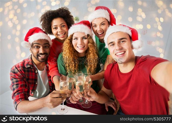 christmas, celebration and holidays concept - happy friends in santa hats clinking glasses of non-alcoholic sparkling wine at home party over lights background. happy friends celebrating christmas at office party