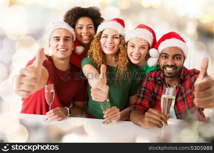 christmas, celebration and holidays concept - happy friends in santa hats with non-alcoholic sparkling wine at party and showing thumbs up over lights background. happy friends showing thumbs up christmas at party