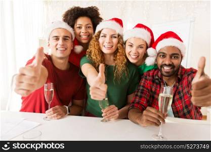 christmas, celebration and holidays concept - happy team in santa hats with non-alcoholic sparkling wine at corporate office party and showing thumbs up. happy team celebrating christmas at office party