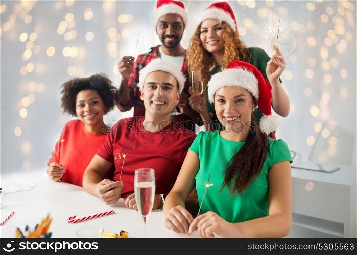 christmas, celebration and holidays concept - happy team in santa hats with sparklers and non-alcoholic sparkling wine at corporate office party over lights background. happy team celebrating christmas at office party