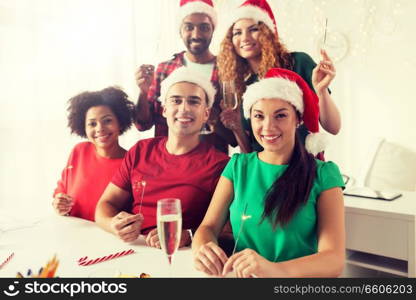 christmas, celebration and holidays concept - happy team in santa hats with sparklers and non-alcoholic sparkling wine at corporate office party. happy team celebrating christmas at office party