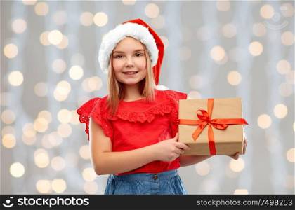 christmas, childhood and holidays concept - smiling girl posing in santa helper hat with gift box over festive lights background. smiling girl in santa hat with christmas gift