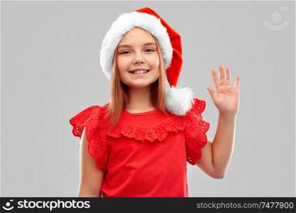 christmas, childhood and holidays concept - smiling girl posing in snata helper hat waving hand over grey background. smiling girl in snata helper hat waving hand