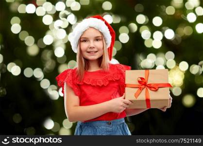 christmas, childhood and holidays concept - smiling girl posing in snata helper hat with gift box over festive lights on dark green background. smiling girl in snata hat with christmas gift