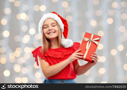 christmas, childhood and holidays concept - smiling pleased girl posing in santa helper hat with gift box over festive lights background. pleased girl in santa hat with christmas gift