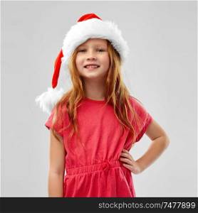 christmas, childhood and holidays concept - smiling red haired girl posing in snata helper hat over grey background. smiling red haired girl posing in snata helper hat