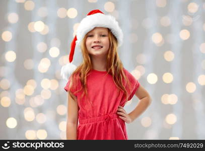 christmas, childhood and holidays concept - smiling red haired girl posing in santa helper hat over festive lights background. smiling red haired girl posing in santa helper hat