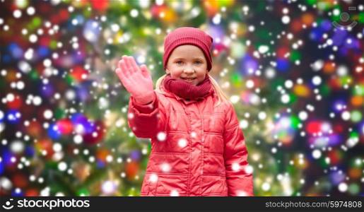 christmas, childhood, gesture, nature and people concept - happy little girl waving hand over snow background and holidays lights