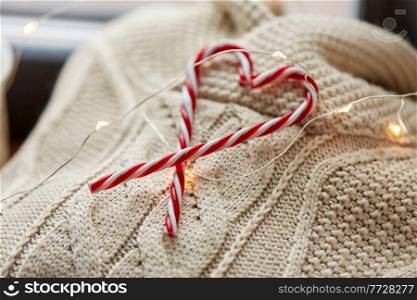 christmas, clothes and objects concept - close up of warm wool braided sweater, candy cane and electric garland string at home. close up of sweater, candy cane, christmas garland