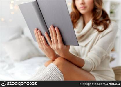 christmas, comfort, leisure and people concept - close up of happy young woman reading book in bed at home bedroom