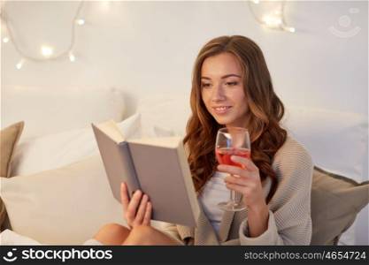 christmas, comfort, leisure and people concept - happy young woman reading book and drinking wine in bed at home bedroom