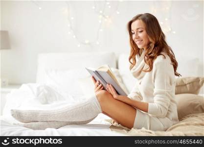 christmas, comfort, leisure and people concept - happy young woman reading book in bed at home bedroom