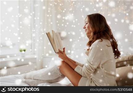 christmas, comfort, leisure and people concept - happy young woman reading book in bed at home bedroom with snow. happy young woman reading book in bed at home