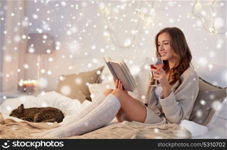 christmas, comfort, leisure and people concept - happy young woman reading book and drinking wine in bed at home bedroom over snow. happy young woman reading book in bed at home