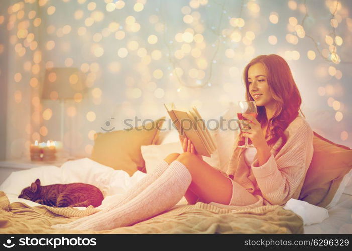 christmas, comfort, leisure and people concept - happy young woman reading book and drinking wine in bed at home bedroom. happy young woman reading book in bed at home