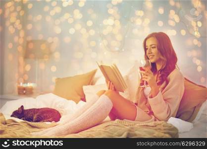 christmas, comfort, leisure and people concept - happy young woman reading book and drinking wine in bed at home bedroom. happy young woman reading book in bed at home