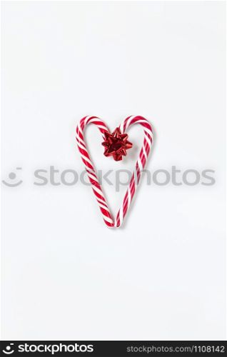 Christmas composition, heart made of two caramel candy canes and red bow on white background. Festive minimal style flat lay. For greeting card, invitation, social media. Vertical orientation.