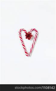 Christmas composition, heart made of two caramel candy canes and red bow on white background. Festive minimal style flat lay. For greeting card, invitation, social media. Vertical orientation.