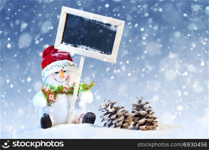 Christmas Concept: Cute Snowman Sitting with Black Board and Pine Cones on the Blue and White Background with Snow