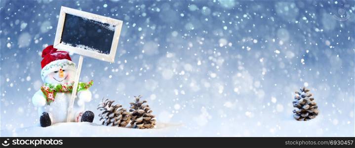 Christmas Concept: Cute Snowman Sitting with Black Board and Pine Cones on the Blue and White Background with Snow