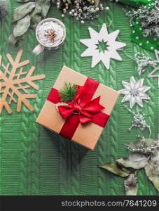 Christmas concept with gift and red ribbon, cup of hot chocolate and snowflakes on green knitted blanket background. Top view. Flat lay