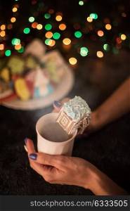 Christmas cookies and cup of tea. Female hands with cup of tea and christmas cookies house on dark color bokeh lights background
