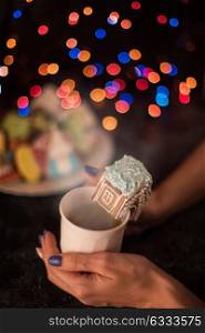 Christmas cookies and cup of tea. Female hands with cup of tea and christmas cookies house on dark color bokeh lights background