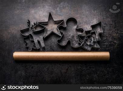 Christmas cookies backing concept. Cookie cutters and baking paper roll on dark rustic background, top view