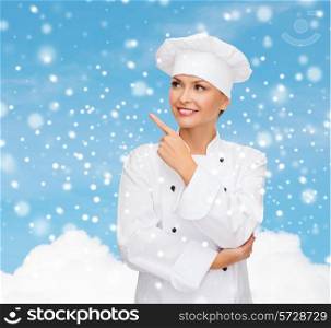 christmas, cooking, holidays and people concept - smiling female chef, cook or baker pointing finger up over blue snowy sky and cloud background