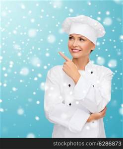 christmas, cooking, holidays and people concept - smiling female chef, cook or baker pointing finger up over blue snowy background