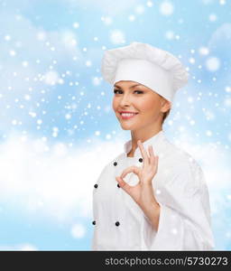 christmas, cooking, profession, gesture and people concept - smiling female chef showing ok hand sign over blue snowy sky and cloud background