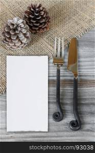 Christmas creative layout composed from steel handmade fork and knife, white paper card and two pine cones are located on the rough wooden table, covered with coarse matting