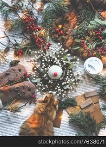 Christmas decoration: fir branches, winter berries, pine cone, cozy mood , fairy lights, paper gift packaging, gloves, coffee and cat around Christmas wreath with snowman. Textile background, Top view