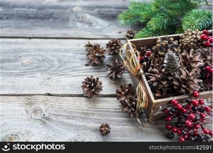 Christmas decoration: full wooden box of pine cones and red holly berries and spruce branches on the background of old unpainted wooden boards; Christmas composition for greeting card, websites, social media, business owners, magazines, bloggers, artists etc.