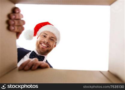 christmas, delivery and winter holidays concept - happy young man in santa helper hat looking into open gift box. happy young man looking into open christmas gift