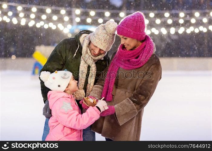 christmas, family and leisure concept - happy mother, father and daughter eating takeaway pancakes at outdoor skating rink in winter. happy family eating pancakes on skating rink