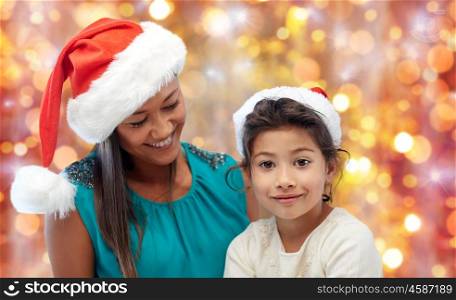 christmas, family, childhood and people concept - happy mother and little girl in santa hats over holidays lights background