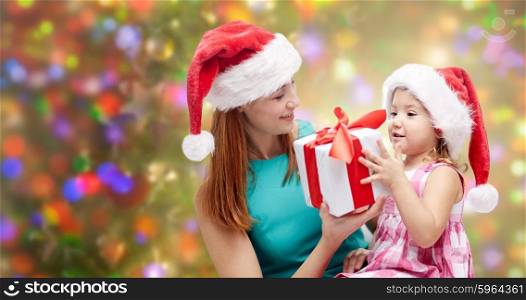 christmas, family, childhood and people concept - happy mother and little girl in santa hats with gift box over holidays lights background